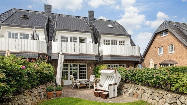 A two-story house with a balcony, a patio with outdoor furniture, and a stone wall surrounding the garden area. - Landhaus Weidenstieg, App. 2 - Luxus und Wohlfühlen am Dorfteich (Wenningstedt-Braderup (Sylt))