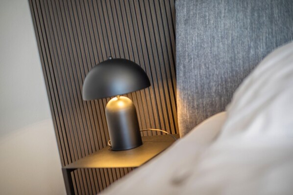 Una mesita de noche moderna con una lámpara, un panel de madera en la pared y una cama blanca.