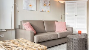 A modern living room with a beige sofa, two checkered pillows, a woven ottoman, a round metal table with a red mug, and two wall-mounted lamps. - Atlantik, App. 10a - Helles Appartement im 1. Obergeschoss, ca. 29 qm Nutzfläche für 2 Personen (Wenningstedt-Braderup (Sylt))
