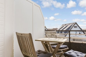Un balcon avec une chaise et une table en bois, un bol en métal, et une vue sur des maisons au loin.