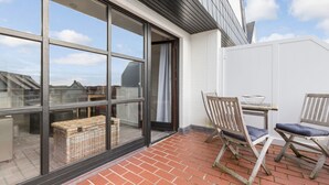 A balcony with a brick floor, two wooden chairs, a wicker basket, and a table with a bowl. - Atlantik, App. 10a - Helles Appartement im 1. Obergeschoss, ca. 29 qm Nutzfläche für 2 Personen (Wenningstedt-Braderup (Sylt))