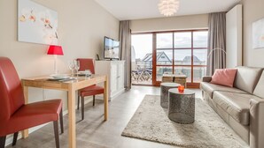 A modern living room with a dining area, a sofa, a TV, and a view of buildings through large windows. - Atlantik, App. 10a - Helles Appartement im 1. Obergeschoss, ca. 29 qm Nutzfläche für 2 Personen (Wenningstedt-Braderup (Sylt))