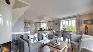 Ein modernes Wohnzimmer mit Kamin, grauen Sofas mit Sternenkissen, einem Holztisch und Blick durch große Fenster auf die Küche und den Essbereich.