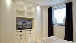 Une chambre avec un téléviseur posé sur un meuble blanc, un lit recouvert de draps blancs, une fenêtre donnant sur une statue et un rideau noir.