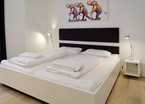 Un lit double avec du linge de lit blanc, une tête de lit noire, des lampes de chevet et un tableau coloré représentant un éléphant accroché au mur.