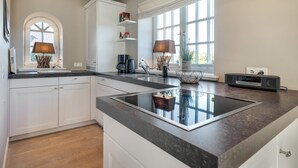A modern kitchen with white cabinetry, a black countertop, and a built-in induction cooktop. There are two pendant lights, a coffee maker, and a radio on the counter. - Sylt-hues - Lifestyle Unter Reet, Strand & Meer um die Ecke. Ihr Optimales Urlaubs-domizil (Wenningstedt-Braderup (Sylt))
