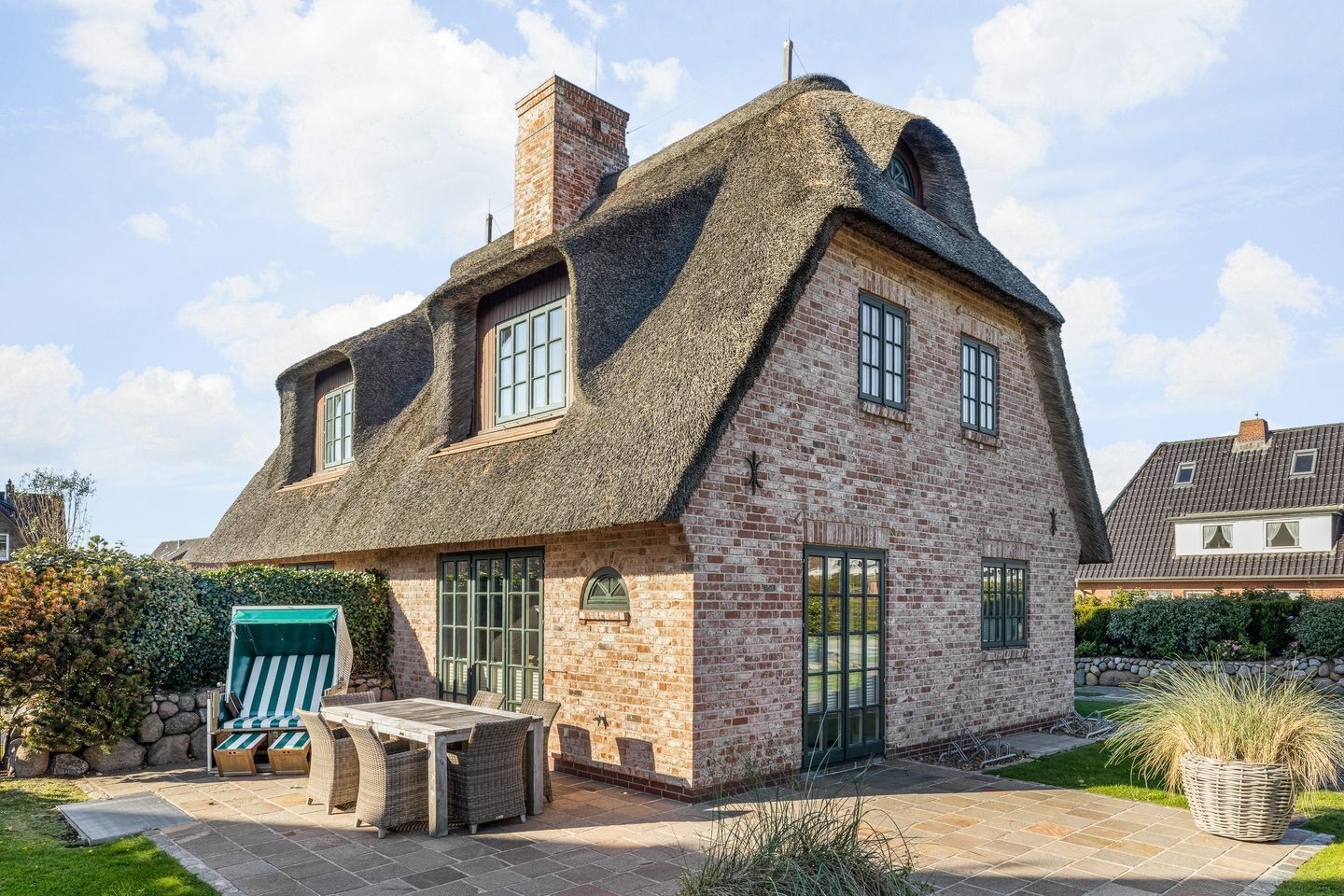 Une maison à deux étages, avec un toit de chaume, des murs en briques et une terrasse aménagée avec du mobilier d’extérieur.