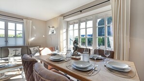 A dining area with a long table set for four, large windows with curtains, a sofa with cushions, and a seahorse decoration. - Sylt-hues - Lifestyle Unter Reet, Strand & Meer um die Ecke. Ihr Optimales Urlaubs-domizil (Wenningstedt-Braderup (Sylt))