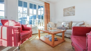 Un salon avec un canapé rouge, une table basse en bois et un canapé beige. De grandes fenêtres dotées de volets bleus offrent une vue sur les bâtiments environnants.