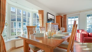 Un coin repas avec une table en bois dressée pour quatre personnes, entourée de chaises, avec un canapé rouge en arrière-plan et de grandes fenêtres offrant une vue sur l’extérieur.