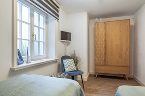 A bedroom with two beds, a window with blinds, a TV, a wardrobe, and a chair with a plant. - Nordic Hüs - Appartement im Eg/ug mit ca. 76 qm Nutzfläche für bis zu 4 Personen (Wenningstedt-Braderup (Sylt))