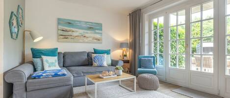 A living room with a grey sofa, a coffee table, and a blue armchair. There is a painting on the wall and a window with curtains.