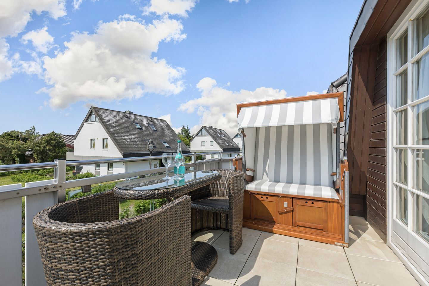 A balcony with wicker furniture, a striped canopy, and a glass table with wine glasses and a bottle.