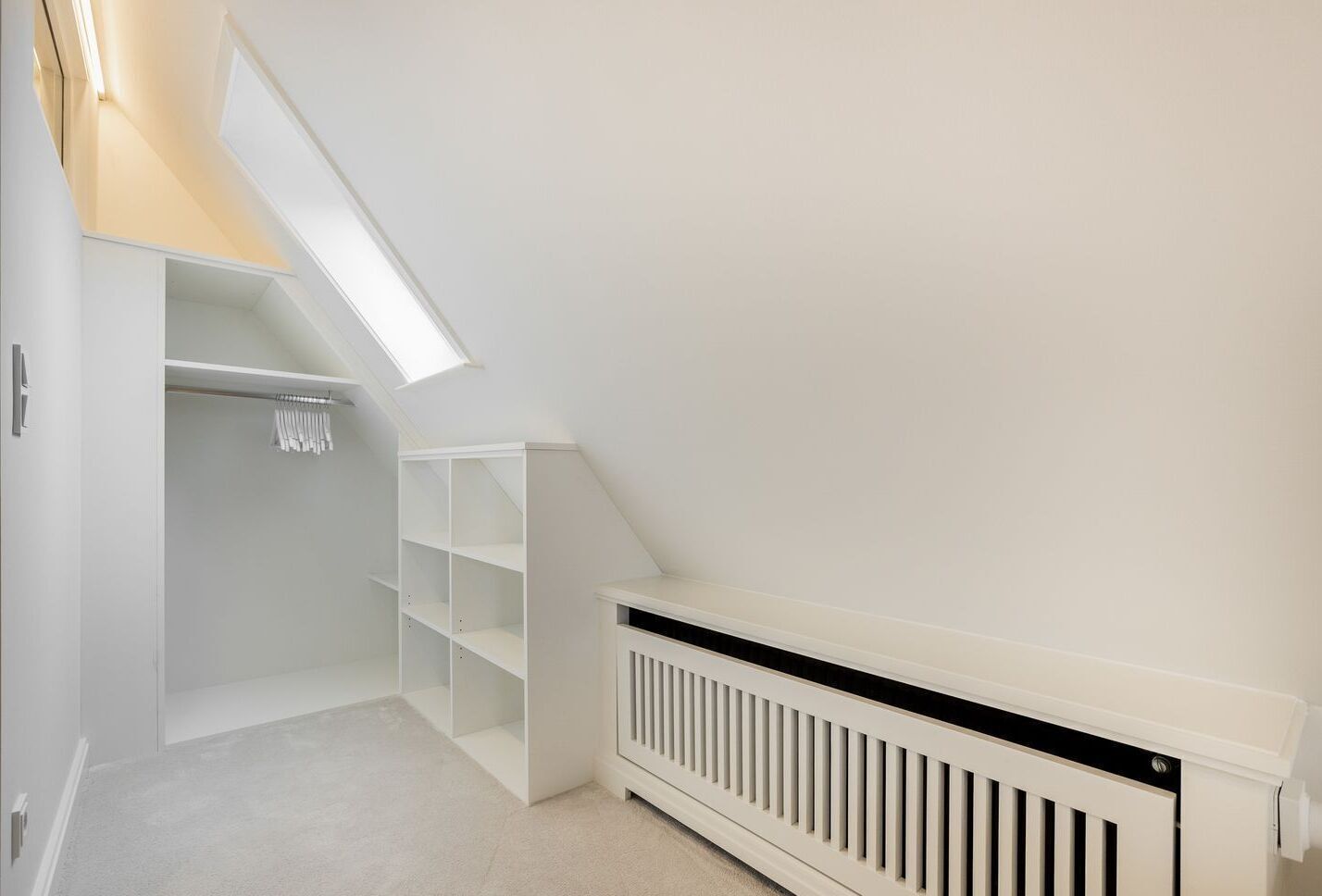 Une chambre spacieuse située dans les combles, avec un plafond en pente, des étagères blanches intégrées, un radiateur et un puits de lumière.