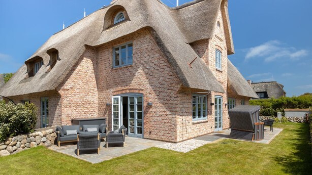 A house with a unique thatched roof and brick walls, a patio with outdoor furniture, and a well-maintained lawn.
