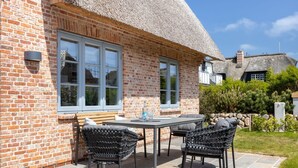 A brick house with a thatched roof, a patio with a table and chairs, and a stone pathway.