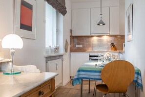 Uma cozinha compacta com armários brancos, uma mesa de jantar com toalha de mesa listrada em azul, uma cadeira de madeira e uma luminária pendente.
