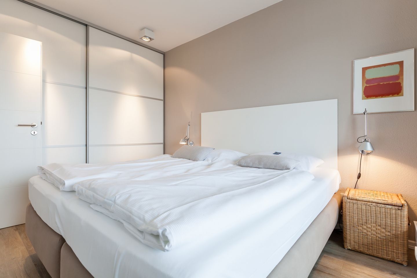 A neatly made bed with white linens, a headboard, bedside lamps, and a wicker basket on the floor.