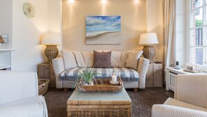 A cozy living room with a sofa, two side tables with lamps, a coffee table with decorative items, and a painting of a beach scene. - Christopher, App. 6 Strönhüs - Urlaubsfeeling Pur. Mit Äußerst Viel Liebe zum Detail (Wenningstedt-Braderup (Sylt))
