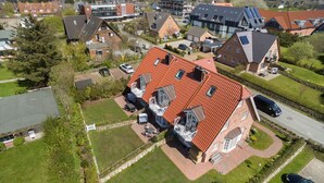 A residential area with a prominent house featuring a red-tiled roof, multiple windows, and a visible outdoor seating area. - Landhaus Normannenweg, App. 4D - In Zentraler und Ruhiger Lage Gelegenes 3-raum Appartement im OG- (Wenningstedt-Braderup (Sylt))