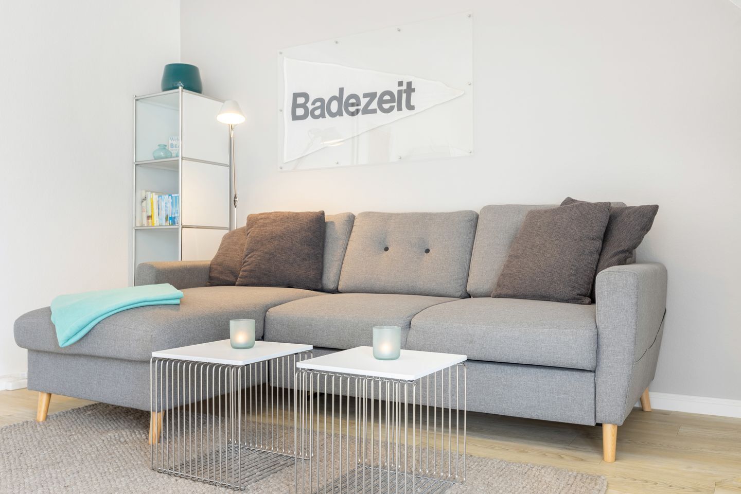 A modern living room with a grey sofa, two white side tables, a metal coffee table, a bookshelf, and a 'Badezeit' sign on the wall.