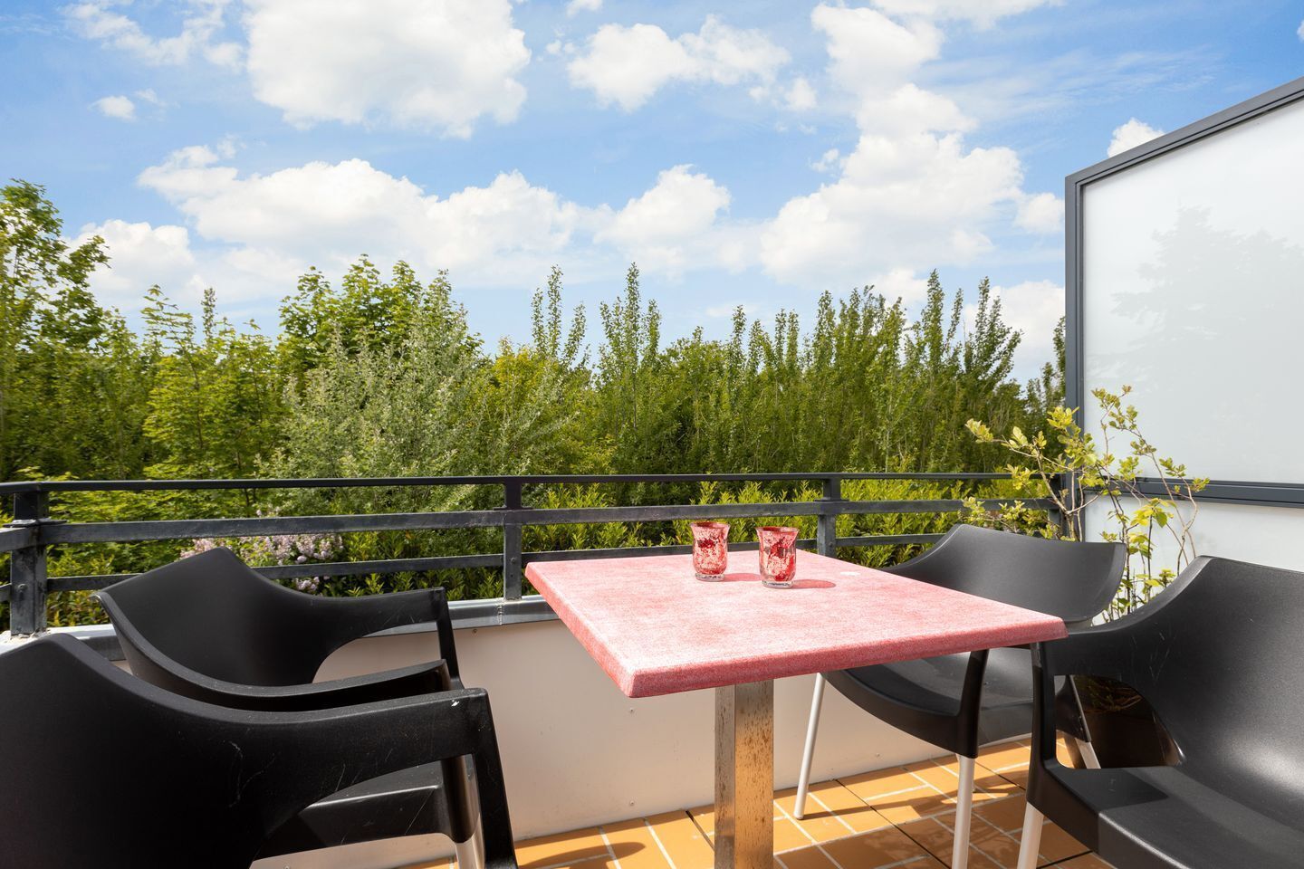 Un balcon avec une table et deux chaises, deux verres contenant une boisson rouge, et une vue sur les arbres et le ciel.