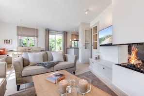 A modern living room with a fireplace, a dining area, and a kitchen visible through an open door.
