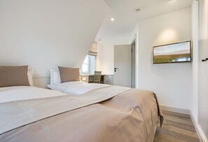 A bedroom with two beds, a desk, and a chair. There is a window with a view of a field and a door leading to another room. - Marysol - Erleben Sie Ihren Urlaub im Typischen Sylt-stil Unter Reet (Wenningstedt-Braderup (Sylt))