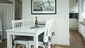 A dining area with a white table and chairs, a framed picture on the wall, and a bottle of wine on the table. - Wallhof, App. 6 - Urlauben und Relaxen im Haus Wallhof - Genießen Sie Sylt in Vollen Zügen (Wenningstedt-Braderup (Sylt))