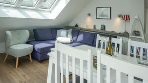 A cozy loft bedroom with a sloped ceiling, a large window, a white bed, a blue sofa, and a white dining table. - Wallhof, App. 6 - Urlauben und Relaxen im Haus Wallhof - Genießen Sie Sylt in Vollen Zügen (Wenningstedt-Braderup (Sylt))