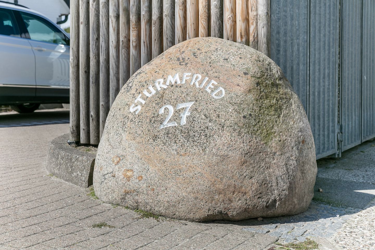 A stone with 'STURMFried 27' engraved on it, placed on a paved surface near a wooden fence and a car.