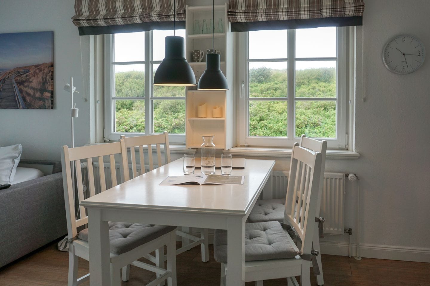 A dining area with a white table, four chairs, a pendant light, and a view of greenery through the window.