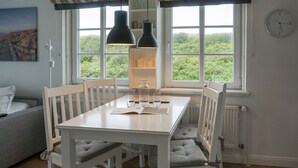 A dining area with a white table, four chairs, a pendant light, and a view of greenery through the window.