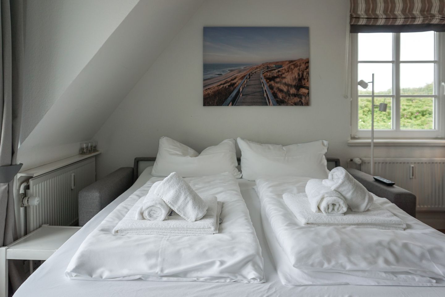 A hotel room with two beds, white linens, a painting of a coastal road, a window with a view of greenery, and a radiator.