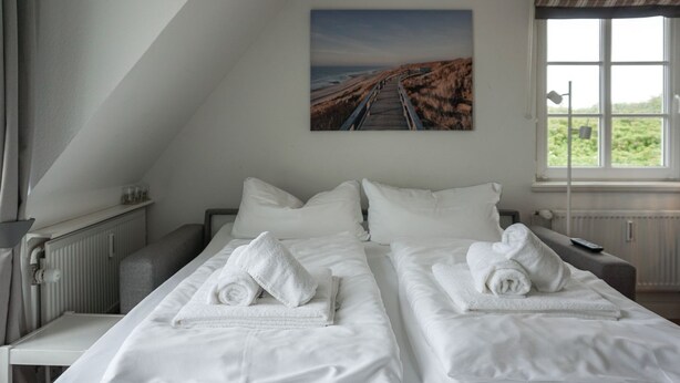 A hotel room with two beds, white linens, a painting of a coastal road, a window with a view of greenery, and a radiator.