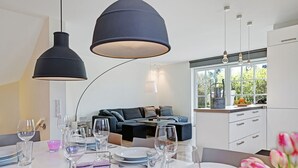 Un comedor moderno con una mesa lista para una comida, un sofá al fondo y una cocina visible a través de una ventana.