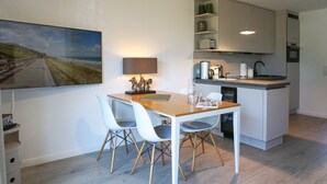 Une cuisine moderne avec une table à manger en bois et des chaises blanches, un îlot central intégré et un tableau représentant une scène de plage.