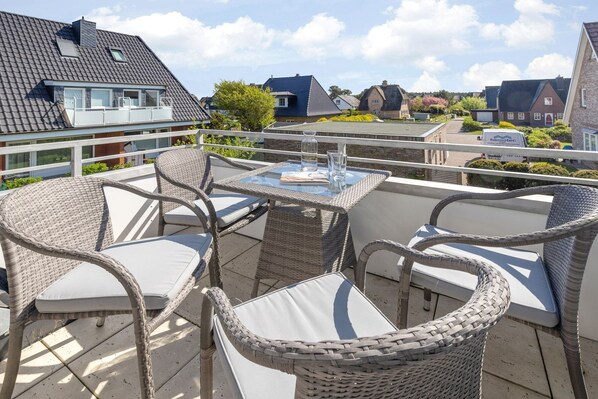 A balcony with wicker furniture and a glass table, overlooking a residential area with houses and a clear sky.