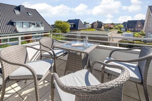 A balcony with wicker furniture and a glass table, overlooking a residential area with houses and a clear sky.