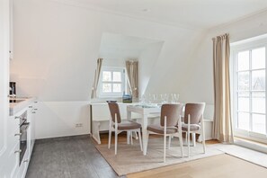 A dining area with a white table set for four, a white chair, a small window with curtains, and a view of the outside. - Westsidehüs - Strand- und Dorfteichnahe Lage. Viel Platz. Eigener Garten. Feine Ausstattung (Wenningstedt-Braderup (Sylt))