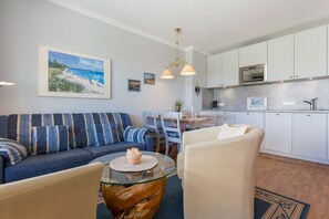 Een moderne woonkamer met een bank, stoelen, een glazen salontafel en een keukenhoek met kastjes en een aanrecht.