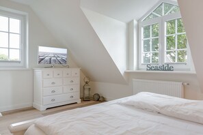 Une chambre avec un plafond en pente, une commode blanche, un lit et une fenêtre donnant sur les arbres.