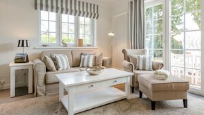 Ein helles Wohnzimmer mit einem beigen Sofa, einem weißen Couchtisch und einem Sessel. Große Fenster mit Vorhängen, eine Stehlampe und ein kleiner Tisch mit Lampe und Büchern sind ebenfalls zu sehen.