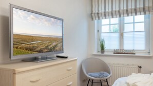 Et soveværelse med et fjernsyn på en kommode, en stol, en seng og et vindue med rullegardiner.