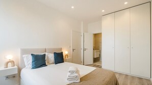 Une chambre moderne avec un grand lit, des lampes de chevet, une armoire et une salle de bain visible à travers une porte ouverte.