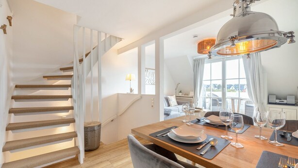 Ein Essbereich mit einem Holztisch, gedeckt für vier Personen, einer Treppe mit weißen Geländern und einem großen Fenster mit Vorhängen.