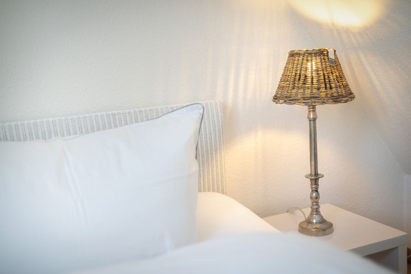 Un lit blanc avec un oreiller blanc et une lampe en métal dotée d’un abat-jour en osier, posée sur une table de chevet blanche.