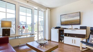 A modern living room with a wooden coffee table, a red sofa, a white cabinet with shelves, and a large window with blinds. - Sylter Refugium, App. 2 - Mitten im Beliebten Inselort Wenningstedt Befindet Sich Dieses Appartement (Wenningstedt-Braderup (Sylt))