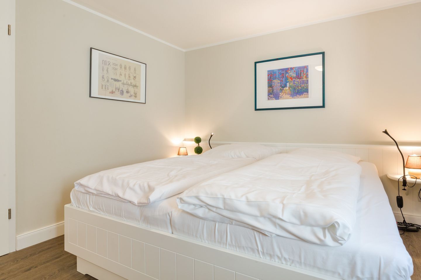 A double bed with white bedding, two wall-mounted lamps, and two framed artworks on the walls.