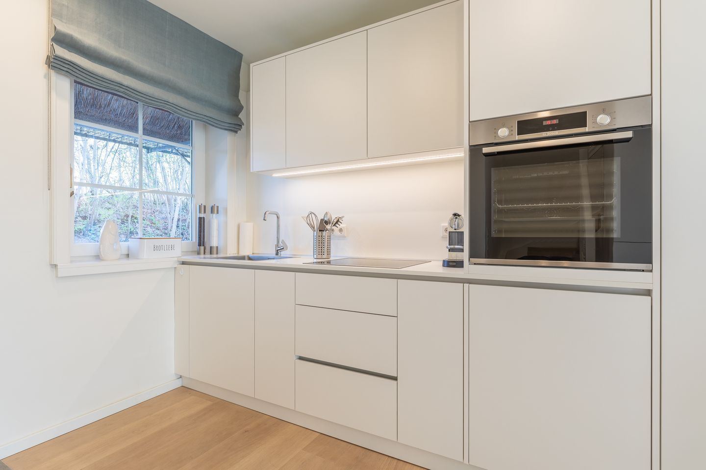 Cocina moderna con armarios blancos, horno integrado y ventana con vista a árboles.